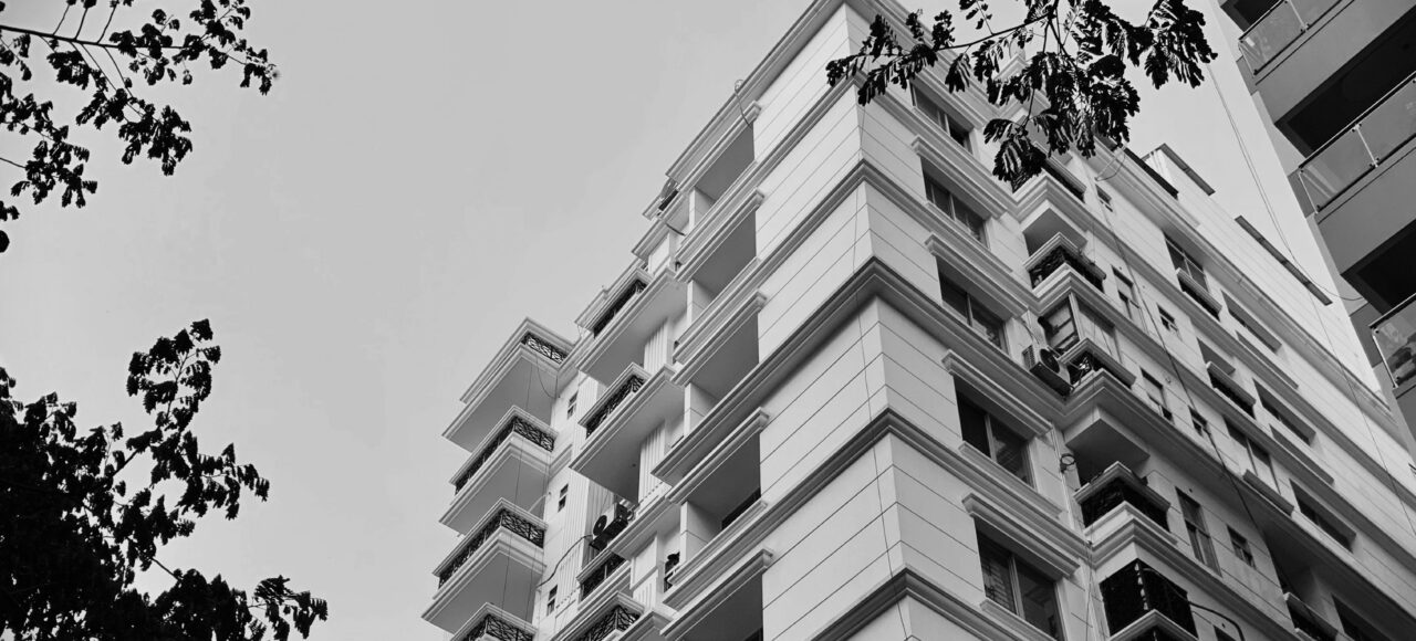 Black-and-white photo of a tall apartment building with multiple balconies viewed from below, framed by tree branches on both sides.